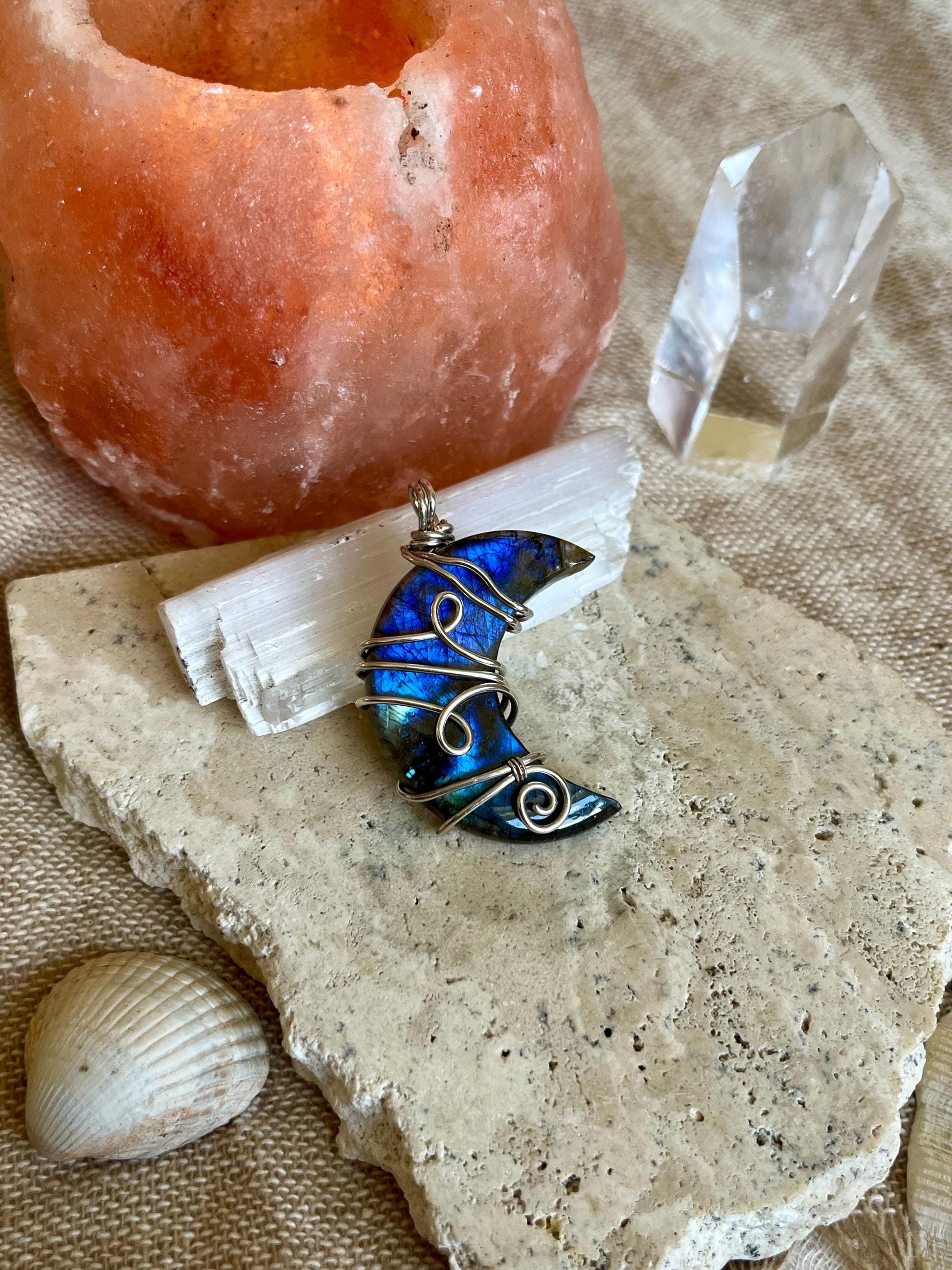 Blue wire-wrapped moon pendant on a stone surface with a crystal and shell.