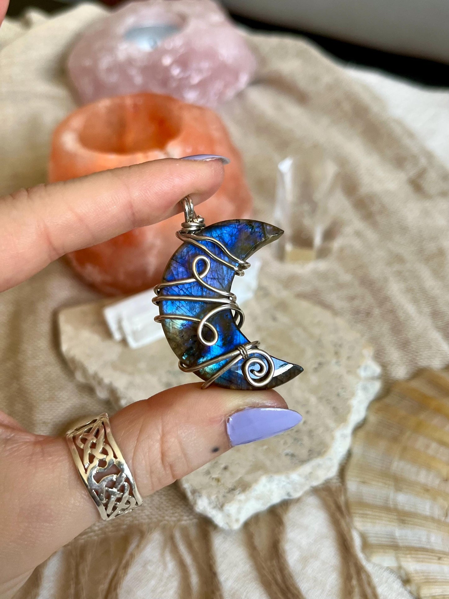 Hand holding a blue moon-shaped pendant with a blurred background of crystals and stones.