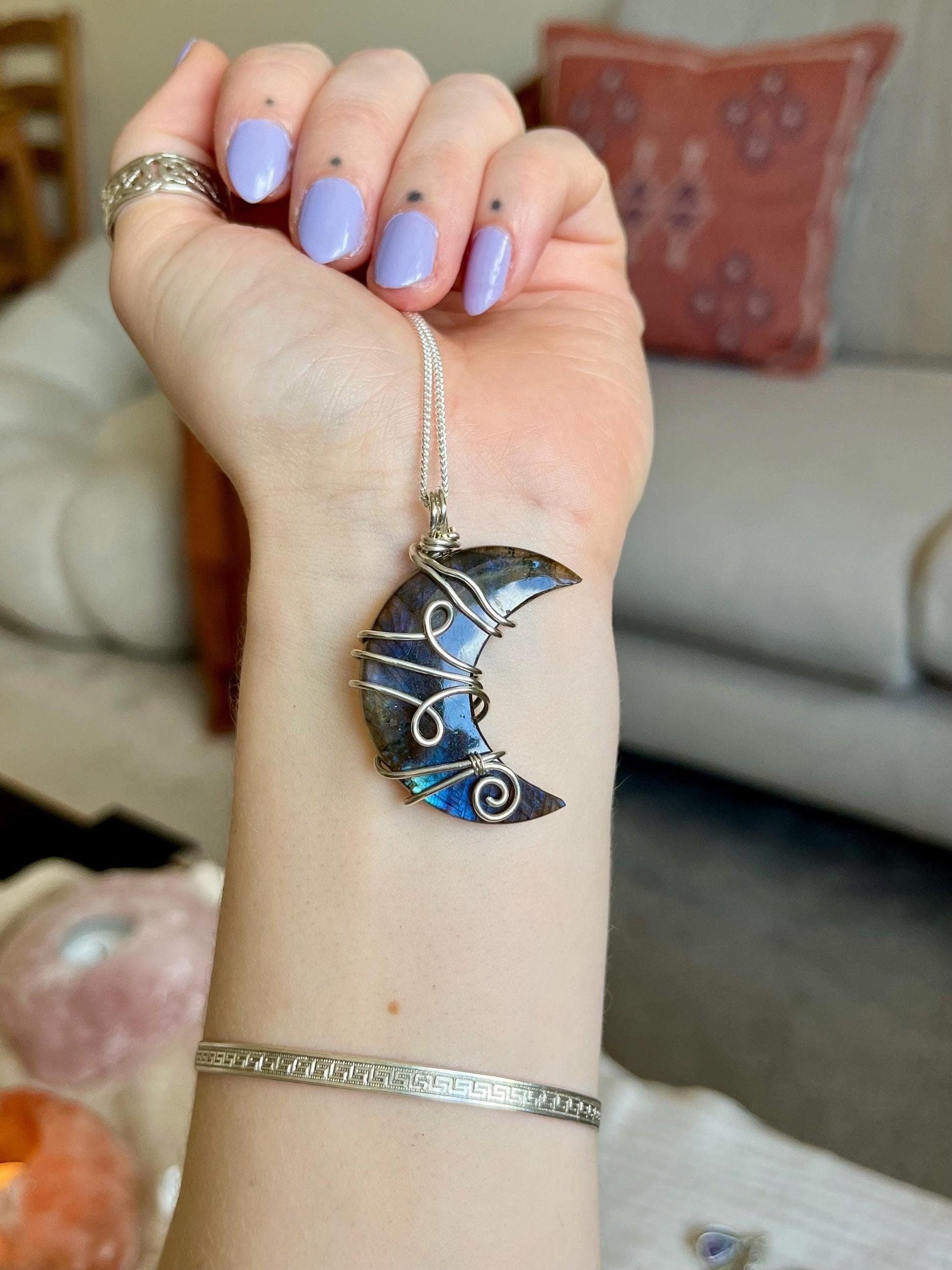 Hand with a silver pendant shaped like a crescent moon with a colorful stone, set against a blurred indoor background.