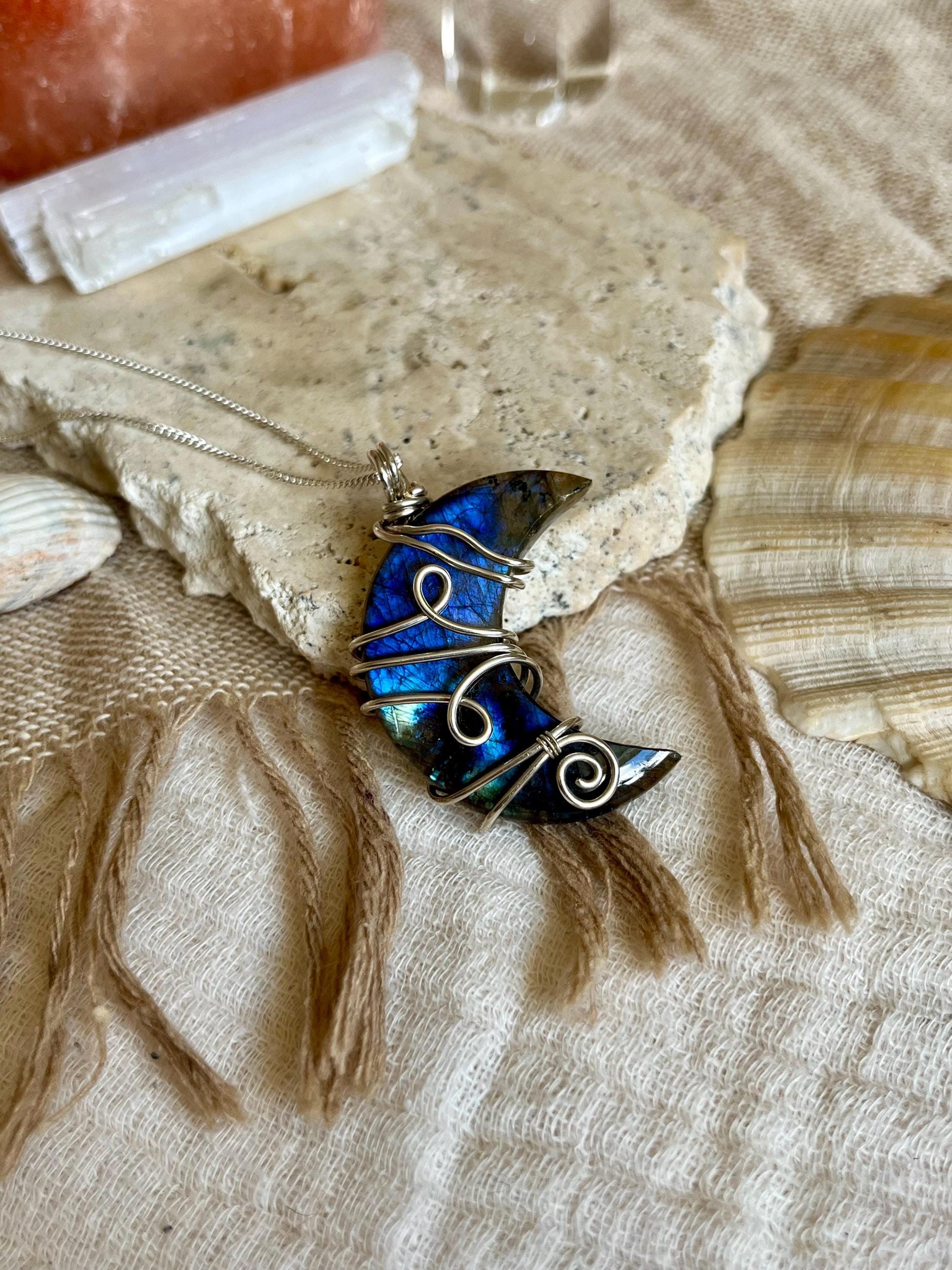 Blue and silver Labradorite pendant on a chain, placed on textured fabric with shells in the background.